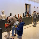 W muzeum ludzie oglądają rzeźby wystawione na ciemnych postumentach. Ściany są białe, podłoga ma jasne, beżowe płyty. Dzieci noszą słuchawki do audioprzewodników.