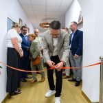 Mężczyzna przecina czerwoną wstęgę w korytarzu z białymi ścianami. Grupa osób w formalnych ubraniach patrzy. Drewniana podłoga.