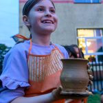 Dziewczynka w pomarańczowym fartuchu trzyma glinianą wazę, uśmiecha się na tle budynku. Jasne ubranie, wieczorne oświetlenie.