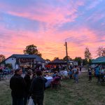 Otwarty festyn z ludźmi na tle różowo-niebieskiego zachodu słońca. Stoiska i scena, wokół zielone drzewa. Atmosfera wieczorna, radosna.