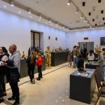 Ludzie zwiedzają galerię pełną rzeźb i popiersi, ściany są białe, podłoga kremowa. Dzieła eksponowane są na ciemnych, prostych cokołach, a sufit oświetlony jest licznymi lampami. W tle w