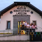 Budynek Muzeum Konstantego Laszczki, o jasnej fasadzie i szarym dachu. Przed wejściem stoi grupa sześciu uśmiechniętych osób. Obok budynku ustawione są kolorowe kosze na śmieci.