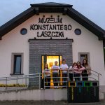 Budynek muzeum w białych i szarych tonacjach z grupą ludzi na tle drzwi. Widoczne kosze na śmieci. W tle wieczorne niebo.