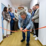 Osoba w dżinsowej kurtce przecina pomarańczową wstążkę w korytarzu. W tle kilka osób na jasnym tle. Na suficie wisi dekoracja.