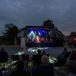 Na zdjęciu jest scena plenerowa z niebieskimi światłami, na której występuje osoba. Widownia siedzi na krzesłach, a w tle widać budynek i ciężarówkę strażacką. Niebieskie i fioletowe reflektory oświetlają scenę