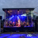 Scena koncertowa na zewnątrz, oświetlona niebieskimi światłami. Na scenie wokalistka i muzycy grają na instrumentach. Tło zasnute chmurami.