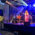 Na scenie w jasnym świetle występuje wokalistka w różowej sukni. Towarzyszy jej zespół muzyczny z bębnami i gitarą. W tle widoczny jest baner z napisem „Mazowsze”.