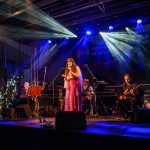 Koncert na scenie; śpiewająca kobieta w różowej sukience, zespół z instrumentami, niebieskie światła w tle.