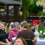 Kilka osób siedzi przy białym stole na zewnątrz, otoczonym zielenią. Na stole jest wazon z kwiatami. W tle widać mężczyznę w koszuli w kratę i zieleni.