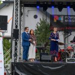 Na scenie przed budynkiem stoją cztery osoby, w tym dwie z mikrofonami. W tle widoczny jest baner Powiat Miński. Na dole widoczny jest napis Mazowsze.