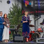 Na scenie cztery osoby; kobieta w niebieskiej sukience mówi do mikrofonu. W tle widoczny bęben i zielone krzewy.