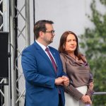 Dwie osoby stoją obok siebie. Mężczyzna w niebieskim garniturze, kobieta w satynowej bluzce, w tle zielone liście. Obok nich widoczna struktura metalowa.