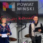 Dwie osoby na scenie z mikrofonami; kobieta w niebieskiej sukience, mężczyzna w kamizelce. W tle baner Powiat Miński.