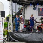 Grupa ludzi stoi na scenie podczas wydarzenia plenerowego. Tło zdobi baner „Powiat Miński”, na scenie są instrumenty muzyczne. Obok sceny stoją głośniki i biały roll-up „Mazowsze”.