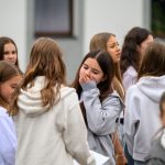 Grupa dziewcząt w jasnych bluzach stoi na zewnątrz, przed szarym budynkiem. Kilka osób rozmawia i śmieje się. Tło jest rozmyte, skupiając uwagę na postaciach.
