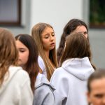 Grupa młodych osób na zewnątrz budynku, w jasnych ubraniach. Dominują odcienie brązu i bieli. W tle szare ściany.