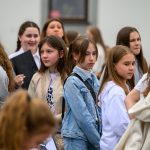 Grupa młodzieży w jasnych ubraniach stoi na zewnątrz. Dominuje kolor biały i szary. Tło jest neutralne, skupione na postaciach.