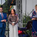 Na zdjęciu widać trzy osoby na scenie: dwie stojące obok siebie i jedną przy mikrofonie. Pierwsza osoba w niebieskim garniturze, druga w ciemnej bluzce i białych spodniach, trzecia w niebieskiej sukni z kwiatami.