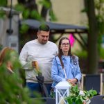 Zdjęcie przedstawia mężczyznę i kobietę na zewnątrz, z zielenią w tle. Mężczyzna trzyma parasol, oboje są w casualowych ubraniach. Kobieta ma ciemne okulary i jasną kurtkę jeansową.