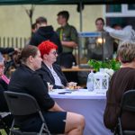 Grupa osób siedzi przy stole z białym obrusem na tle namiotu. Na stole są kwiaty i filiżanki. W tle stoją inni ludzie i budynek.