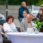 Na zdjęciu widać ludzi przy stole z białym obrusem i zielonym bukietem. Tło tworzą niebieskie i szare detale zewnętrzne. Każda osoba przyjmuje różne pozy i zajęcia.