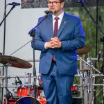 Mężczyzna w niebieskim garniturze przemawia na scenie. Za nim znajduje się zestaw perkusyjny i transparent z napisem. Scena oświetlona, w tle drzewa.