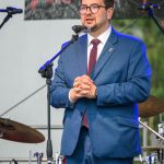 Mężczyzna w niebieskim garniturze przemawia na scenie. W tle znajdują się perkusja i napis w czerwieni. Scena jest oświetlona światłem dziennym.