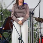 Na zdjęciu kobieta w szarej bluzce i białych spodniach stoi przy mikrofonie. Tło jest neutralne, z elementami sprzętu muzycznego. Scena ma delikatne zielone akcenty.