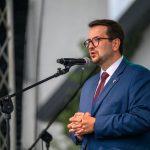 Mężczyzna w niebieskim garniturze przemawia do mikrofonu. Scena na świeżym powietrzu, w tle metalowe rusztowanie. Wygląda formalnie.