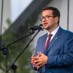 Mężczyzna w niebieskim garniturze przemawia do mikrofonu. Ma okulary i ciemne włosy. Tło jest rozmyte, z zielenią i szarościami.
