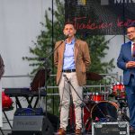 Na scenie stoi trzech ludzi z mikrofonami. W tle widać sprzęt muzyczny i zielone liście. Mężczyzna w środku mówi do mikrofonu.
