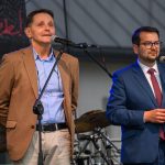 Dwóch mężczyzn stoi przed mikrofonami na scenie. Jeden ma brązową marynarkę, drugi niebieski garnitur. W tle widać instrumenty muzyczne.