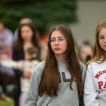 Grupa młodych osób stoi na zewnątrz. Ubrani są w sportowe bluzy, w tle widać rozmazane postacie. Tło jest zielone, nadając wrażenie parku.