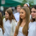 Na zdjęciu widać kilka młodych osób na zewnątrz, ubranych na biało. Tło jest rozmyte z zielonymi i ciemnymi elementami. Dominują naturalne kolory i subtelne uśmiechy.