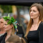 Na zdjęciu są dwie kobiety, jedna w wianku z liści, druga w czarnej sukience. W tle rozmyta grupa ludzi. Dzień jest pochmurny.