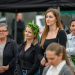 Grupa ludzi stoi na zewnątrz. Kobieta ma wianek z liści. W tle są namioty w ciemnych kolorach.