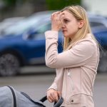 Osoba w jasnym płaszczu stoi na ulicy z wózkiem, poprawiając włosy. W tle widoczny jest rozmyty, niebieski samochód. Obraz ma neutralne, miejskie otoczenie.