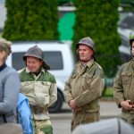 Obraz przedstawia trzech mężczyzn w zielono-beżowych mundurach z hełmami, stojących na zewnątrz. W tle widać zielone drzewa i biały samochód. Na pierwszym planie kobieta z dzieckiem.