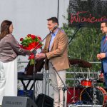 Trzy osoby na scenie, kobieta wręcza bukiet czerwonych róż mężczyźnie. Tło to sprzęt muzyczny i zielone drzewa. Strój elegancki.