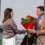 Mężczyzna w brązowej marynarce wręcza bukiet czerwonych róż kobiecie na scenie. Tło jest jasne, z fragmentami zieleni. Obok znajduje się mikrofon na statywie.
