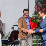 Trzech ludzi na scenie; mężczyzna w garniturze podaje dłoń drugiemu, trzymającemu czerwone róże. Kobieta patrzy z boku.