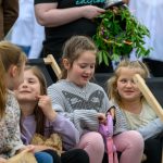 Grupa uśmiechniętych dzieci siedzi na leżakach na zewnątrz. Dziewczynki rozmawiają w swobodnej atmosferze. W tle widać osoby dorosłe i zielony wianek.