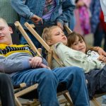 Dzieci odpoczywają na leżakach. Chłopiec w żółtej koszulce, dziewczynki w zielonej i białej. Tło jest rozmyte i wielobarwne.