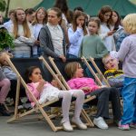 Dzieci siedzą na leżakach w kolorowych ubraniach, otoczone grupą rówieśników. W tle zielono-beżowy namiot. Scena na świeżym powietrzu.