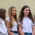 Trzy dziewczyny w białych koszulkach stoją na zewnątrz, na tle jasnej ściany. Przeważa kolor biały i beżowy. W tle widoczne są rozmyte postacie.