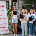 Grupa dzieci trzyma zdjęcia Piłsudskiego, stojąc przy banerze Mazowsze. Ubrane w jasne ubrania, śpiewają na zewnątrz. W tle rośliny i scena.