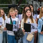 Grupa dzieci w białych bluzkach i dżinsach śpiewa na scenie, trzymając czarne teczki. W tle widoczne instrumenty perkusyjne. Dzieci mają przed sobą zdjęcia mężczyzny z brodą.