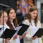 Grupa młodych ludzi śpiewa z nutami na scenie, ubrani w białe stroje. Tło czarne, mikrofon z przodu. Skupienie na twarzach.
