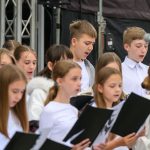 Grupa dzieci w białych strojach śpiewa, trzymając czarne teczki. Tło techniczne, metalowe konstrukcje sceny. Dzieci skupione, mikrofon przed nimi.