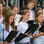 Grupa dzieci śpiewa, trzymając czarne teczki z nutami. Ubrane są w białe bluzki i stoją na tle mikrofonów. Scena ma neutralne, ciemne tło.